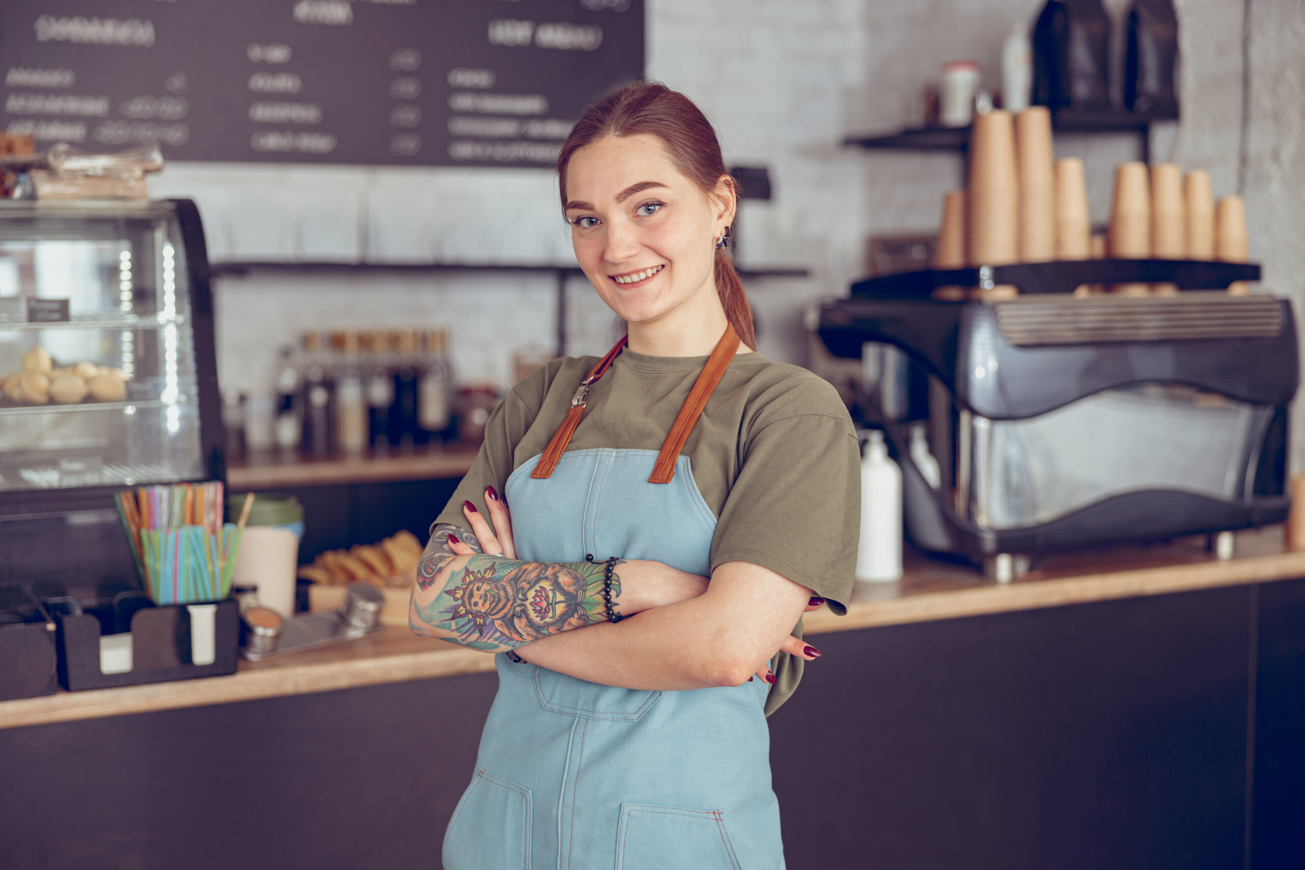 Joyful young woman cafe worker looking at camera and smiling while crossing arms over her chest